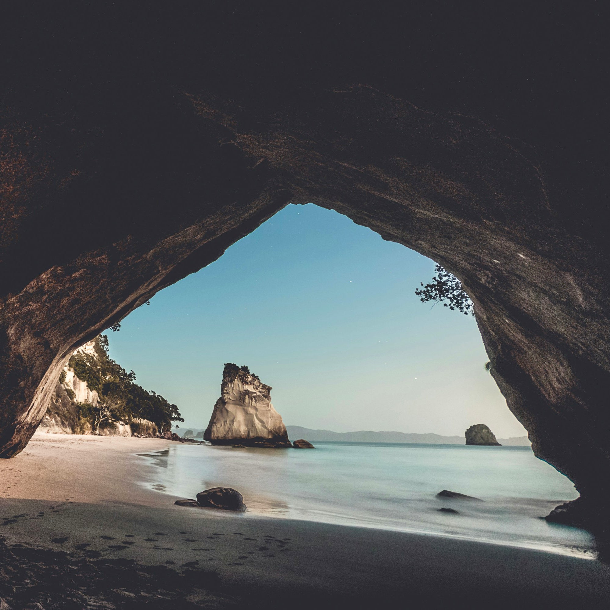 A scenic photograph of a New Zealand beach viewed through a large natural rock arch, with pale sand, calm blue water, distant rock formations, and a clear sky.