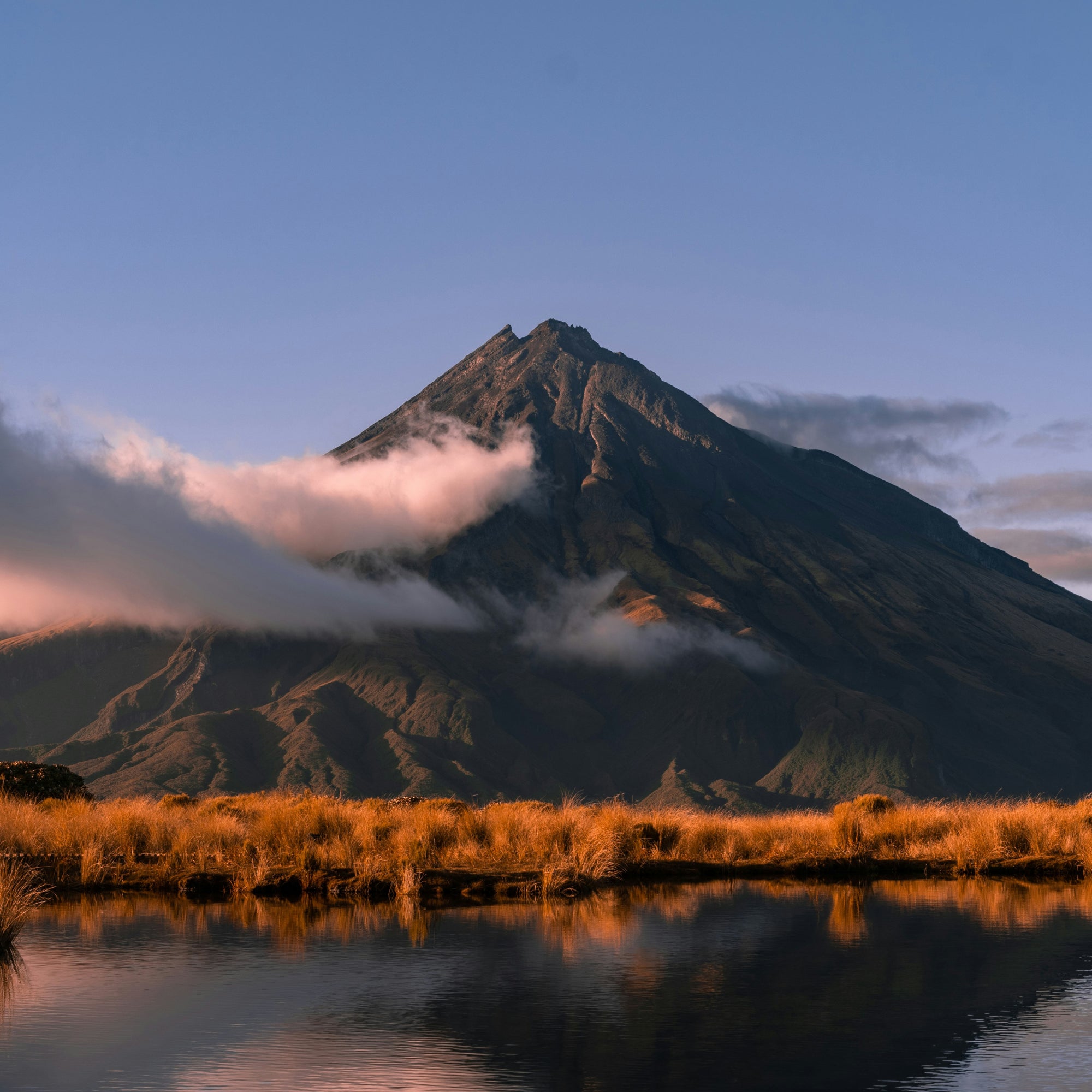A scenic landscape photograph of the Mt Taranaki mountain peak rising above golden tussock grasses and calm water, with low clouds drifting around the slopes under a clear blue sky.