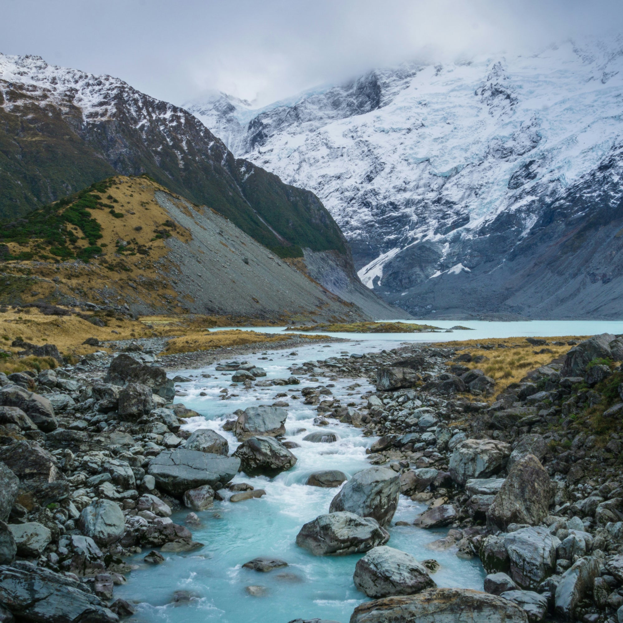 A dramatic mountain landscape in New Zealand Southern Alps with snow-covered peaks, a rocky stream with turquoise water flowing through the foreground, and a pale blue lake in the distance under an overcast sky.
