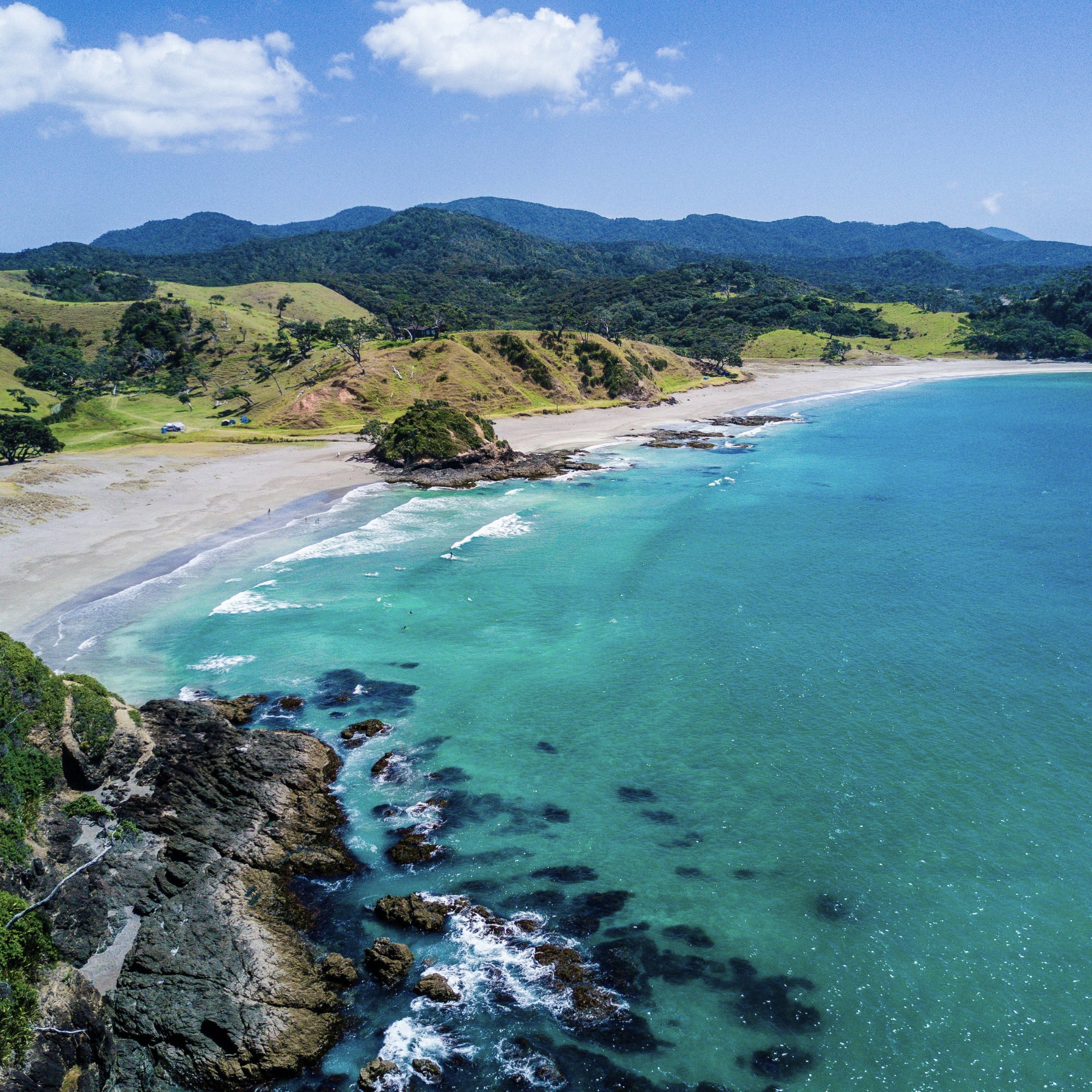 A white sandy beach with rock formations between grassy hills covered in native NZ bush, and a calm blue sea in the bay.