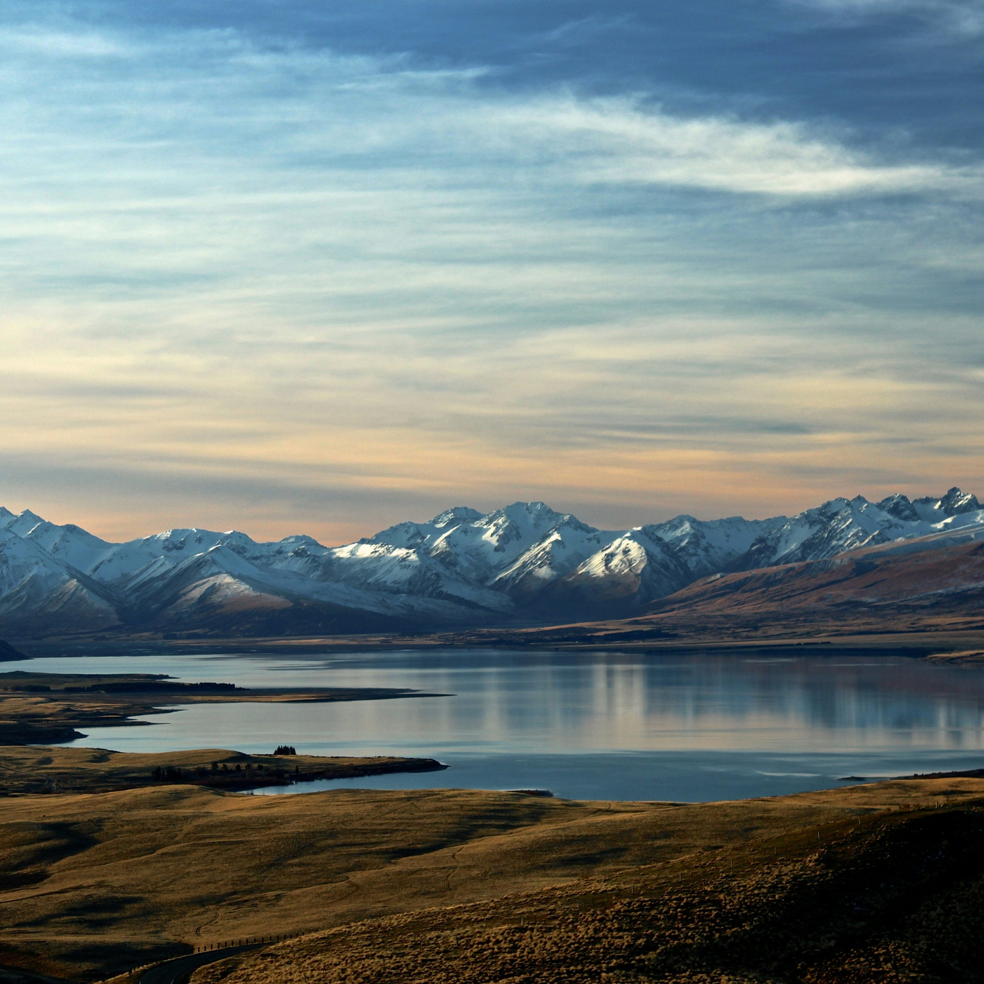 A scenic landscape photograph of a calm lake in New Zealand reflecting snow-capped mountains under a blue sky with wispy clouds.