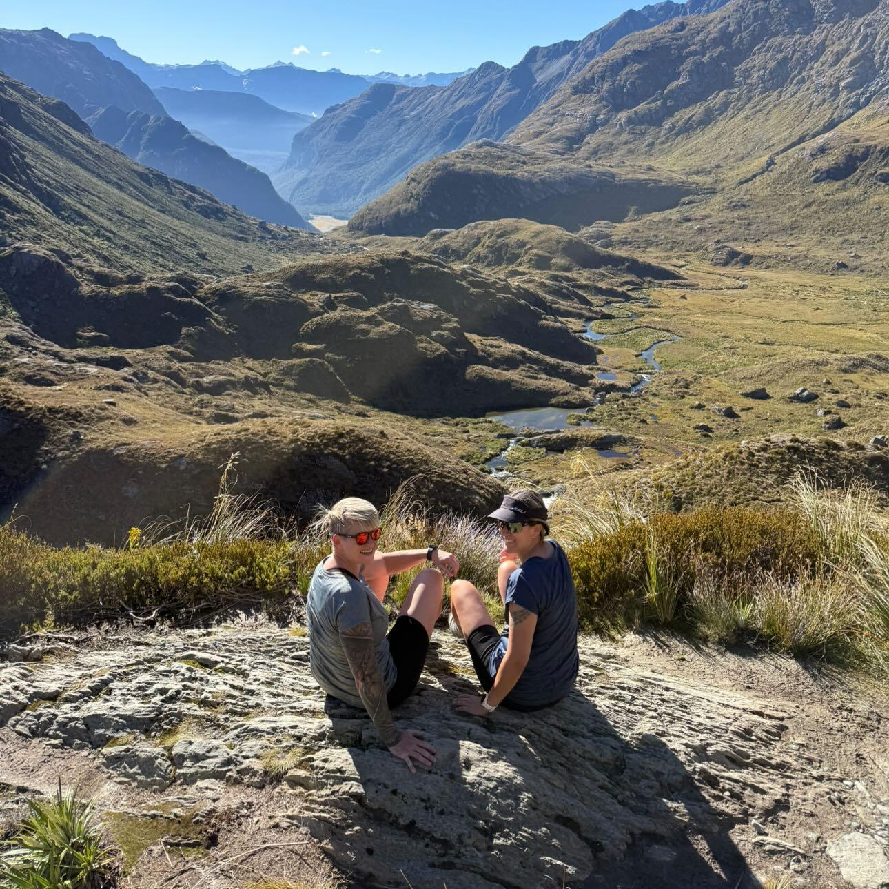 Earthing NZ owners Kelly & Jess pose next to a blue lake at the top of a grassy valley in the New Zealand highlands with the sun beaming down.