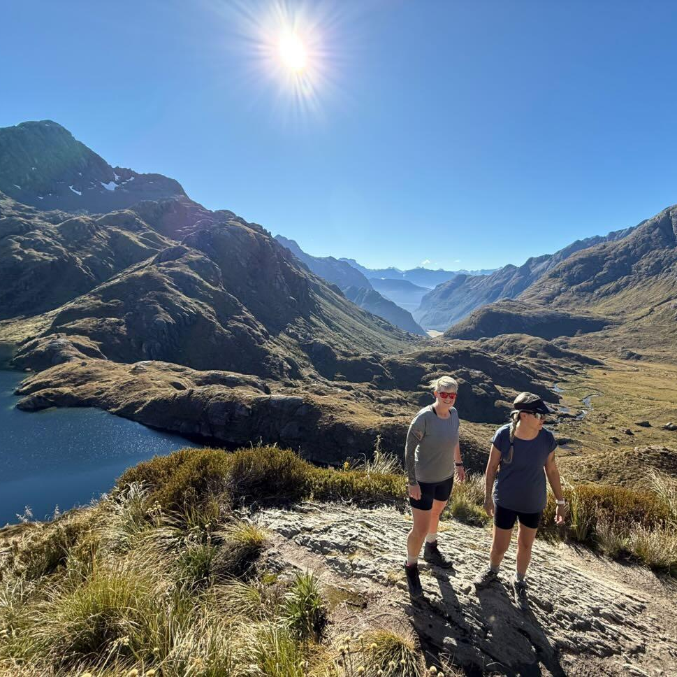 Earthing NZ owners Kelly & Jess pose next to a blue lake at the top of a grassy valley in the New Zealand highlands with the sun beaming down.